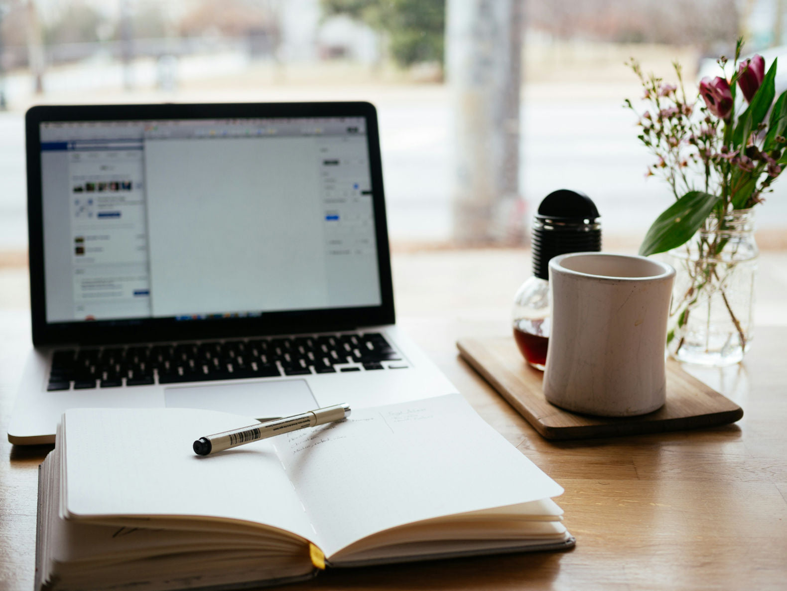 A cozy workspace with a laptop, notebook, pen, coffee, and wooden gifts.